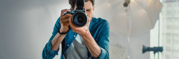 In the Photo Studio with Professional Equipment: Portrait of the Famous Photographer Holding State of the Art Camera Taking Pictures with Softboxes Flashing in Background.